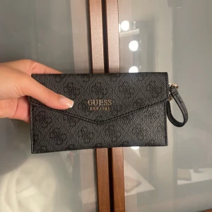 Svart plånbok från Guess - Snygg svart plånbok från Guess med diskret logomönster och guldfärgad logga framtill. Plånboken har en praktisk handledsrem och är tillverkad i syntetmaterial med struktur. Perfekt för att hålla koll på kort och kontanter.