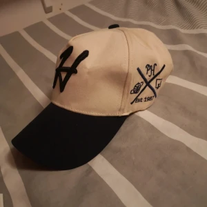 Beige och marinblå keps PH 1987 - Snygg keps i beige med marinblå skärm och broderade detaljer. Framsidan har ett stort svart 'H' och sidan är dekorerad med blå brodyr, texten 'PH', 'Est 1987' och coola symboler. Justerbar baktill med metallspänne. Perfekt accessoar för en sportig look.