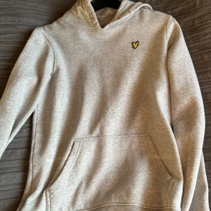 Grå hoodie från Lyle & Scott - Säljer en stilren grå hoodie från Lyle & Scott med klassisk gul logga på bröstet. Tröjan har huva, känguruficka och långa ärmar. Perfekt för dig som gillar en enkel och clean look. Jätte skönt och varmt material.
