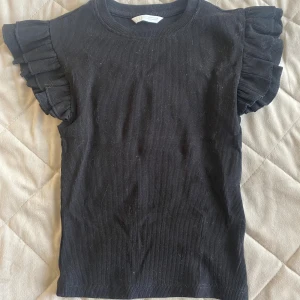 Svart ribbad topp med volangärm Zara - Svart ribbad topp från Zara med volangdetaljer på axlarna. Toppen är ärmlös och har en rund halsringning. Materialet är mjukt och stretchigt, perfekt för en trendig look. Snygg att styla med jeans eller kjol.