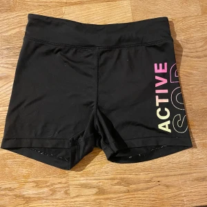 Svarta sportshorts från H&M Sport - Svarta, tighta sportshorts från H&M Sport med färgglad text 'ACTIVE' i rosa, gul och lila på ena benet. Har en vit fläck på rumpan