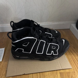 Nike Air More Uptempo svarta sneakers - Svarta Nike Air More Uptempo sneakers med feta vita AIR-bokstäver på sidorna och klassisk Swoosh-logga. Skorna har snörning, platt sula och en chunky siluett. Ovandelen är i mocka och syntet, och de har en riktigt sportig vibe som sticker ut.