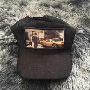 Svart keps från Maggiore med mesh - Svart keps från Maggiore med meshpaneler på sidorna och justerbar rem bak. Framtill finns en patch med bild på en gul sportbil och en man i kostym. Kepsen har en metallogo på sidan och insidan är märkt med Maggiore-logga.