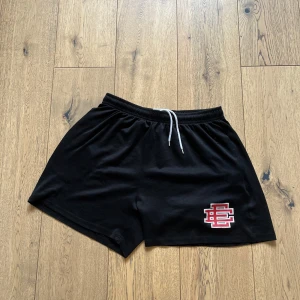 Svarta shorts från Eric Emanuel - Svarta shorts från Eric Emanuel med vit snörning i midjan och EE-logga i rött och vitt på vänster ben. De har elastisk midja och är tillverkade i ett mjukt, sportigt material som passar perfekt för chill eller träning.
