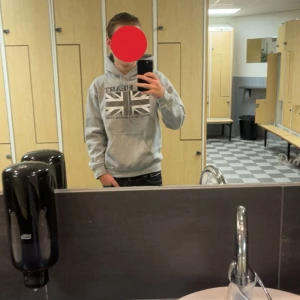 Grå hoodie Gildan med brittiskt tryck - Grå hoodie från Gildan med tryck av brittiska flaggan och texten 'MY HEART BEATS TO A BRITISH TUNE' på bröstet. Klassisk känguruficka, snörning i huvan och mjukt material. Perfekt för dig som gillar streetstyle och vill sticka ut med ett coolt statement.