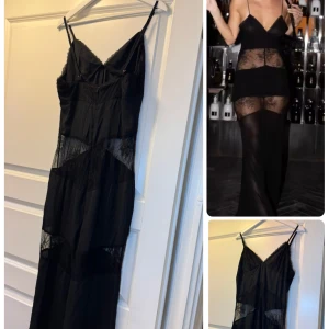 Svart långklänning med spetsdetaljer - Svart långklänning med tunna axelband och v-ringning. Klänningen har genomskinliga partier i spets som ger en cool och edgy look. Figurnära passform och slits för extra attityd. Perfekt för dig som vill sticka ut med en dramatisk och trendig stil.