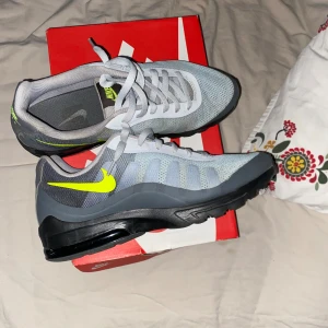 Nike Air Max Invigor grå sneakers - Nike Air Max Invigor sneakers i grå mesh med svarta detaljer och en neon-gul swoosh. Skorna har en luftig ovandel, synlig Air Max-dämpning i sulan och klassisk snörning. Perfekta för dig som gillar sportig och modern stil.