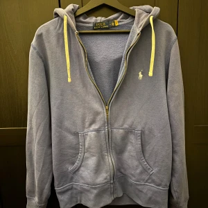 Blå hoodie från Polo Ralph Lauren - Blå hoodie från Polo Ralph Lauren med vit snörning och dragkedja framtill. Klassisk broderad logga på bröstet och två fickor. Mjuk insida och huva för extra chill vibe. Perfekt för dig som vill ha en clean och stilren look.