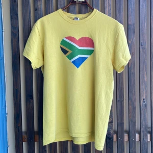 Gul t-shirt Sydafrika - Gul t-shirt från Gildan med tryck av ett hjärta i Sydafrikas flagga på bröstet och texten 'SIGMA WOW TOUR' på ryggen. På ärmen finns en liten färgglad stjärna och texten 'Star for Life'. Skön bomullskvalitet och klassisk passform.