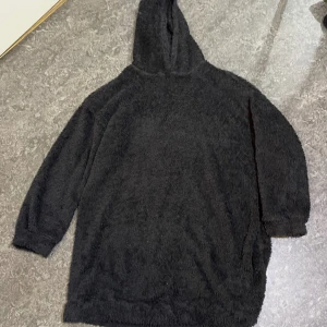 Svart fluffig hoodie med huva - Mysig svart hoodie i fluffigt material med huva och långa ärmar. Perfekt för kalla dagar när du vill ha något riktigt bekvämt och oversized. Enkel design utan tryck eller detaljer, bara ren komfort.