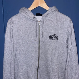 Grå Patagonia hoodie med dragkedja - Grå zip-hoodie från Patagonia med svart logga på bröstet och stort tryck på ryggen. Tröjan har huva med snörning, två fickor fram och är tillverkad i mjuk bomullsmix.