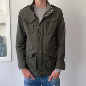 Olivgrön fieldjacket från J.Lindeberg - Snygg olivgrön vindjacka från J.Lindeberg med fyra stora fickor framtill, axelklaffar och dragkedja med tryckknappar. Jackan har en clean look och är tillverkad i lätt polyester, perfekt för vår och höst. Sann i storleken och passar alltså medium. Jag har inga mått fråga inte efter det. Kom med bud i DM.