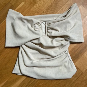 Beige offshoulder topp med rynk - Trendig beige topp med offshoulder-design och breda ärmar. Toppen har snygga rynkade detaljer framtill och är tillverkad i ett mjukt material som ger en skön passform. Perfekt för dig som vill sticka ut med en cool och modern look.