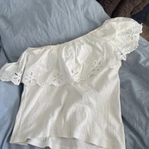 Vit offshoulder topp med volang från ZARA  - Superfin vit offshoulder topp med bred volangkant och broderade blommor. Toppen är ribbad och har en luftig känsla, perfekt för varma dagar. Volangen har snygga hålbroderier som ger en romantisk vibe. Size 13-14, cm 164