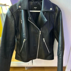 Svart skinnjacka från Bik Bok XS - Snygg svart skinnjacka från Bik Bok i bikerstil med silvriga dragkedjor och tryckknappar. Jackan har klassisk krage, flera fickor med dragkedja och en croppad passform. Perfekt för dig som vill ha en edgy look.