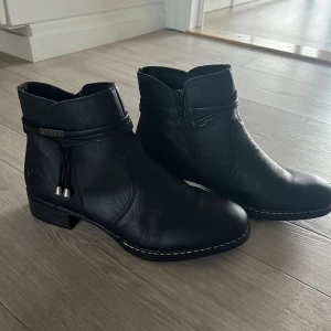 Svarta boots från Rieker med dragkedja - Snygga svarta boots från Rieker i skinn med mjukt foder på insidan. Skorna har rund tå, låg klack och dragkedja på sidan. Dekorativa detaljer runt skaftet och robust platt sula med Rieker-logga undertill. Aldrig använda endast testade