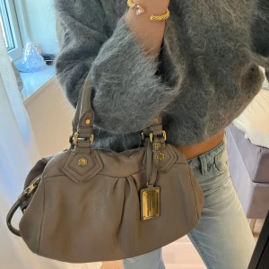 Snyggaste beigea Q Classic Baby Groove väskan!  - Snyggaste beigea handväskan i äkta läder från Marc by Marc Jacobs. Den ikoniska modellen Q Classic Baby Groove! En populär och eftertraktad vintagemodell med gulddetaljer och dubbla handtag. Väskan är i fint skick. Helhetsintrycket är mycket välbevarat.  Det här är en tidlös modell som inte längre finns att köpa, vilket gör den extra eftertraktad. Perfekt för dig som vill ha en stilren tidlös väska med riktigt bra andrahandsvärde. Passar lika bra till vardag som till fest.  Mått: 34×18×12 cm 