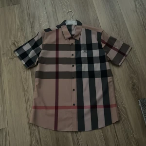 Rutig kortärmad skjorta Burberry XXL - Burberry skjorta i klassiskt rutigt mönster med beige, svart, röd och gråa toner. Skjortan har korta ärmar, knappar framtill och en snygg krage. Perfekt för dig som gillar statement-plagg och vill sticka ut med en ikonisk design.