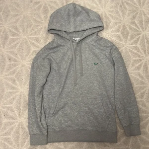 Lacoste hoodie - Säljer en stilren grå hoodie från Lacoste i 100% bomull. Tröjan har huva med snörning, lång ärm och den klassiska gröna krokodilloggan på bröstet. Regular fit och mjukt material som är perfekt för chill dagar.