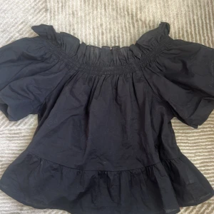 Svart offshoulder topp Samsa Collection - Supersöt svart offshoulder topp från Samsa Collection med volangkant upptill och nertill. Toppen har korta puffärmar och smockad resår runt axlarna för en snygg passform. Perfekt att styla med jeans eller kjol för en trendig look.