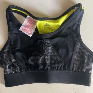 Svart och gul linnetopp från Adidas - Sportig linnetopp från Adidas i svart och neon-gul med coolt leopardmönster på sidorna och loggan. Tillverkad i polyester med AEROREADY-teknologi för extra komfort vid träning. Perfekt för dig som gillar att sticka ut på gymmet eller idrotten.