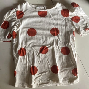 Vit t-shirt med röda prickar Minimarket - Vit t-shirt från Minimarket med stora röda prickar och puffiga korta ärmar. Tillverkad i mjuk bomull, har en rund halsringning och en avslappnad passform. Perfekt för dig som gillar lekfulla prints och coola detaljer.