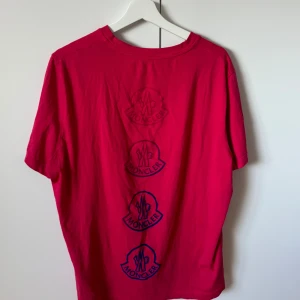 Rosa Moncler t-shirt med logga - Snygg rosa t-shirt från Moncler i mjuk bomull. T-shirten har korta ärmar och flera Moncler-loggor tryckta vertikalt både fram och bak. Passformen är normal.