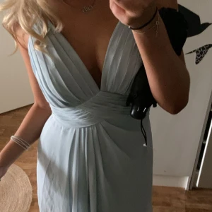 Ljusblå balklänning med spetsrygg - Supersnygg ljusblå lång balklänning med v-ringad omlott framtill, smala axelband och slits på sidan. Baksidan har transparent spetsdetalj och knappar. Klänningen har ett flowigt fall och markerad midja, perfekt för dig som vill sticka ut med en elegant look.