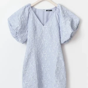 Ljusblå Brocade klänning från Gina Tricot - Superfin ljusblå eller silveraktig klänning med puffärmar och v-ringning från Gina Tricot. Klänningen har ett strukturerat, blommigt mönster och är tillverkad i ett syntetmaterial som ger en cool textur. Passformen är normal och klänningen slutar ovanför knäna. Perfekt för alla tillfällen!