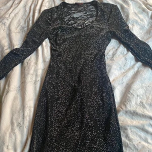 Svart glittrig klänning med spets - Supersnygg svart klänning med glittriga detaljer och långärmade ärmar. Baksidan har en stor spetspanel med blommigt mönster som ger en cool kontrast. Klänningen är figurnära och har en v-ringad halsringning framtill. Perfekt för dig som vill sticka ut med lite extra glitter.En super fin fest/ ny års klänning😍 + 60kr Frakt