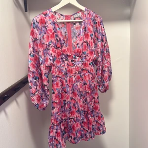 Blommig kortklänning från Loavies XS - Superfin kortklänning från Loavies i färgstarkt blommigt mönster med rosa, lila och röda toner. Klänningen har v-ringning, volangkant nertill och långa puffärmar. Perfekt för dig som gillar färg och vill sticka ut lite extra!