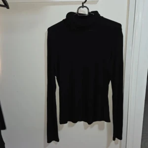 Svart polotröja från H&M, storlek M - En svart polotröja från H&M i storlek M. Tröjan har lång ärm och en klassisk hög polokrage. Den är figurnära och tillverkad i ett mjukt, stretchigt material som känns skönt mot huden. Perfekt att styla med jeans eller kjol.