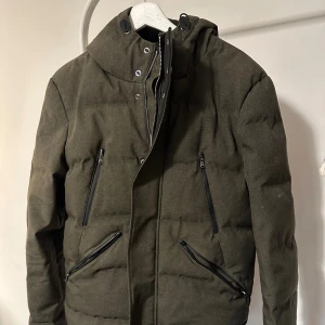 Olivgrön pufferjacka från IKKS Urban Lab - Snygg olivgrön pufferjacka från IKKS Urban Lab med fyra dragkedjeförsedda fickor och hög krage med tryckknappar. Jackan har en modern look och är perfekt för kyliga dagar. Materialet är polyester och jackan har en stilren, mörkgrön ton. Det finns lite smuts på armen i första bilden men jag har tagit bort det nu. :)
