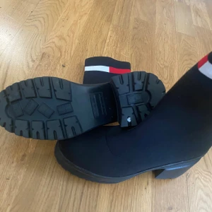 Svarta stickade boots med klack - Snygga svarta boots från Tommy Hilfiger med stickad ovandel och breda ribbade muddar upptill i rött, vitt och marinblått. Grov sula och stabil klack ger en cool look. Perfekta för dig som vill ha både stil och edge.Aldrig använts 