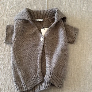 Beige stickad kofta från Zara - Kortärmad stickad kofta från Zara i beige nyans. Koftan har en bred krage, ribbade muddar och stängs med en knapp framtill. 