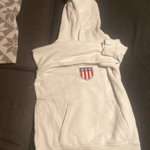 Vit gant hoodie med amerikanskt emblem - Vit hoodie med huva och känguruficka framtill. På bröstet sitter ett emblem med amerikanska flaggans färger och stjärnor. Perfekt för dig som gillar enkel stil med en touch av USA. Mjuk och skön, passar till många outfits.