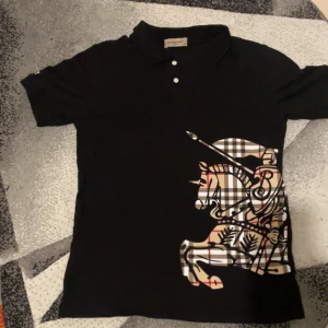 Pike från ett fint märke  - Svart t-shirt från fint märke med klassisk krage och tre knappar framtill. Stor rutig print med riddare och häst i beige, svart, vitt och rött på framsidan. Broderad -logga på ärmen. Snygg och stilren design med exklusiv känsla.