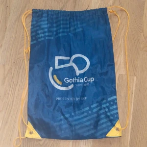 Blå gympapåse Gothia Cup - Säljer en blå gympapåse från Gothia Cup med gult snöre och tryckt logga. Väskan har dragsko och är perfekt för träning eller att bära med sig småsaker. Lätt och smidig att ta med överallt.