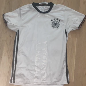Tysklands vit fotbollströja - Vit fotbollströja från Tysklands landslag med svarta detaljer och broderat landslagsemblem på bröstet. Tröjan har svarta ränder längs sidorna, svart krage och korta ärmar med diskreta ränder i tysklands färger. Tillverkad i glansigt, lätt polyestermaterial.