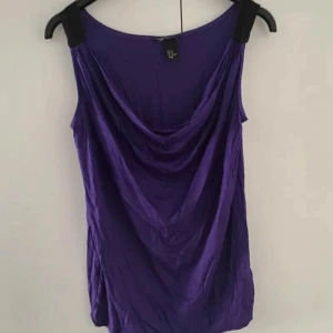 Lila draperad linne från H&M - Säljer en snygg lila linnetopp från H&M med draperad urringning och svarta axelband. Toppen är ärmlös. Sparsamt använd