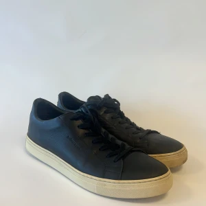 Svarta sneakers från Björn Borg - Stilrena svarta sneakers från Björn Borg med klassisk låg profil och vit platt sula. Skorna är tillverkade i skinn och har snörning framtill. Perfekta för dig som gillar minimalistisk design och vill ha ett par schyssta sneakers till vardags.