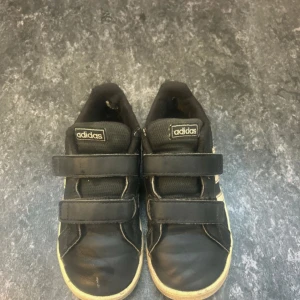 Svarta Adidas sneakers med kardborre - Svarta sneakers från Adidas med klassiska vita ränder på sidan och vit sula. Skorna har två praktiska kardborreband för enkel på- och avtagning. Rund tå och platt sula ger en skön och avslappnad stil. Perfekt för dig som gillar sportig vibe och enkel design.