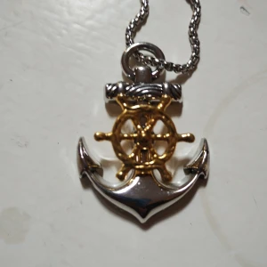 Halsband med ankare och ratt i silver och guld - Unikt halsband med ett detaljerat hänge i form av ett ankare och ett skeppsratt. Hänget är i silverfärg med gulddetaljer på ratten och sitter på en klassisk silverfärgad kedja. Perfekt accessoar för dig som gillar marina vibbar och snygga detaljer.