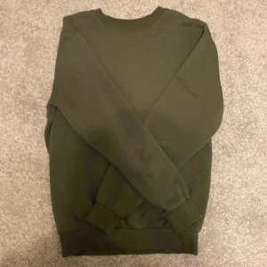 Mörkgrön crewneck sweatshirt - En basic mörkgrön crewneck sweatshirt med rund halsringning och ribbade muddar vid ärmslut och nederkant. Tröjan är enkel och stilren, perfekt att matcha med jeans eller joggers. Materialet känns mjukt och bekvämt mot huden.