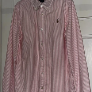 Rosa randig skjorta från Ralph Lauren - Klassisk rosa och vit randig skjorta från Ralph Lauren med broderad logga på bröstet. Skjortan har lång ärm, knappar hela vägen och button down-krage. Perfekt för en preppy look och tillverkad i mjuk bomull.