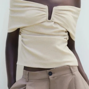 Beige offshoulder topp från Zara - Trendig beige offshoulder topp från Zara med snygg v-ringning och rynkade detaljer framtill. Toppen är croppad och har en figurnära passform som framhäver siluetten. Perfekt att styla med höga byxor för en cool look.