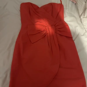 Rosa  axelbandslös klänning H&M 38 - Snygg rosa axelbandslös kortklänning från H&M i storlek 38. Klänningen har en stor rosett framtill och omlott-detalj vid bysten. Perfekt för dig som vill sticka ut med en färgstark look. Materialet är mjukt och följsamt. Klänningen ser röd ut på bilden men den är rosa