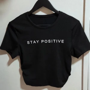 Svart croppad topp med text, M - Svart croppad topp med vit text 'STAY POSITIVE' framtill. Rund halsringning och korta ärmar. T-shirten är tight i passformen och tillverkad i mjukt bomullsmaterial. Perfekt för dig som gillar enkel och clean stil med ett peppigt budskap.