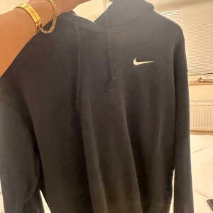 Svart Nike hoodie med huva - Svart hoodie från Nike med klassisk vit logga på bröstet. Tröjan har huva med snörning och långa ärmar. Perfekt för chill dagar eller när du vill ha en sportig look.
