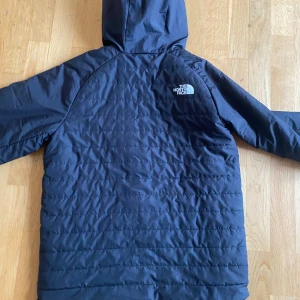 Svart pufferjacka från The North Face - Snygg svart pufferjacka från The North Face med huva och dragkedja framtill. Jackan har diskret quiltad yta, logga på bröstet och ärmen, samt två sidofickor med dragkedja. Perfekt för kyliga dagar och enkel att matcha med allt.
